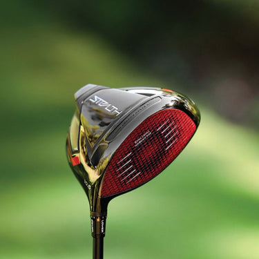 Taylormade stealth driver being held up with a blurred green background. The stealth drivers red face is showing with a view of the black sole as well. 